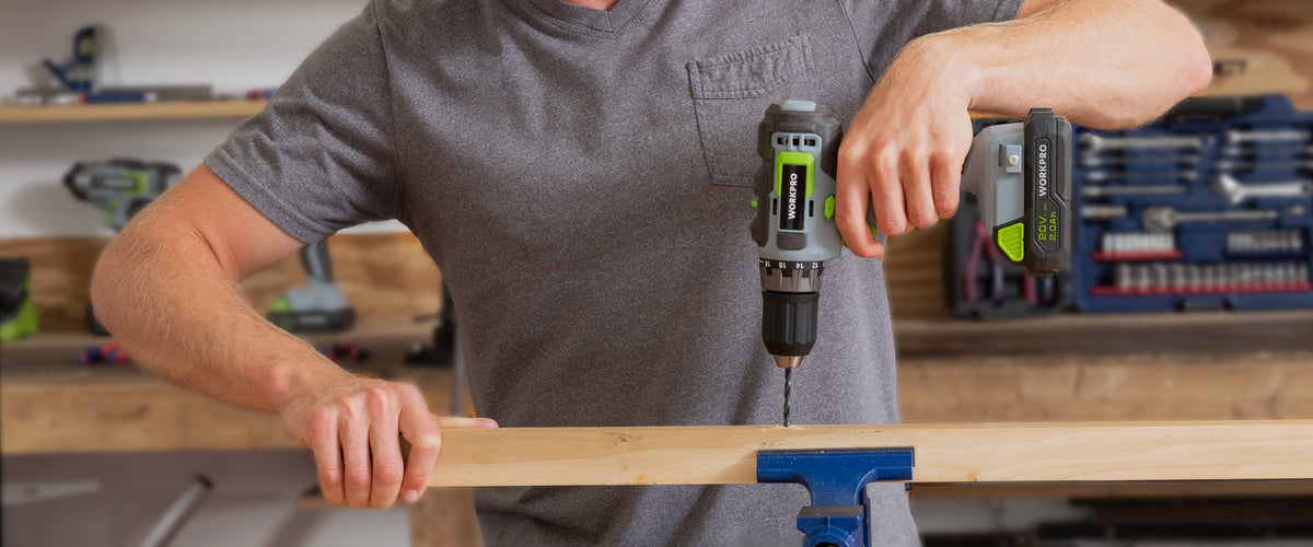 Person using a cordless drill to fasten a wooden board held in a blue bench vise in a workshop