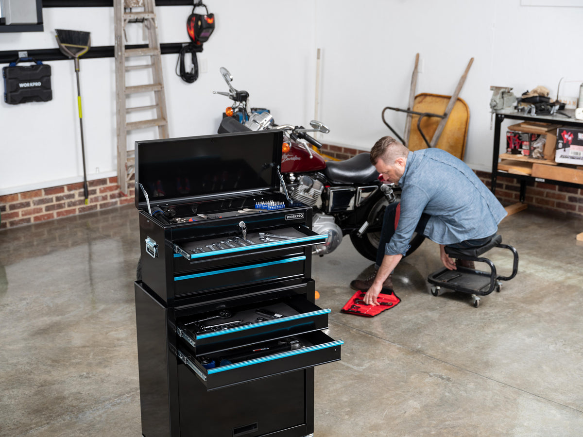 Black rolling tool chest with open drawers beside a motorcycle, while a worker kneels on a garage floor