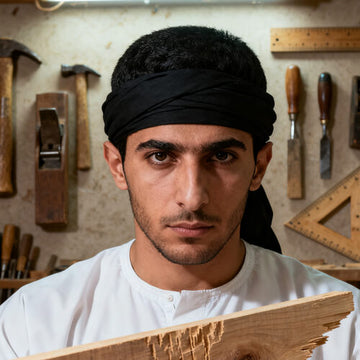 Carpenter wearing a black headband and white shirt, holding a wooden plank in a workshop with tools
