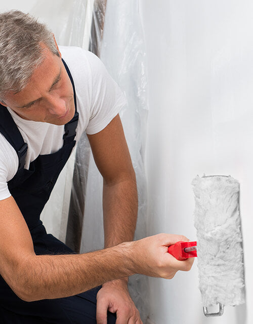 A painter in a white shirt and dark apron applying white roller paint to a wall, showing innovation and quality