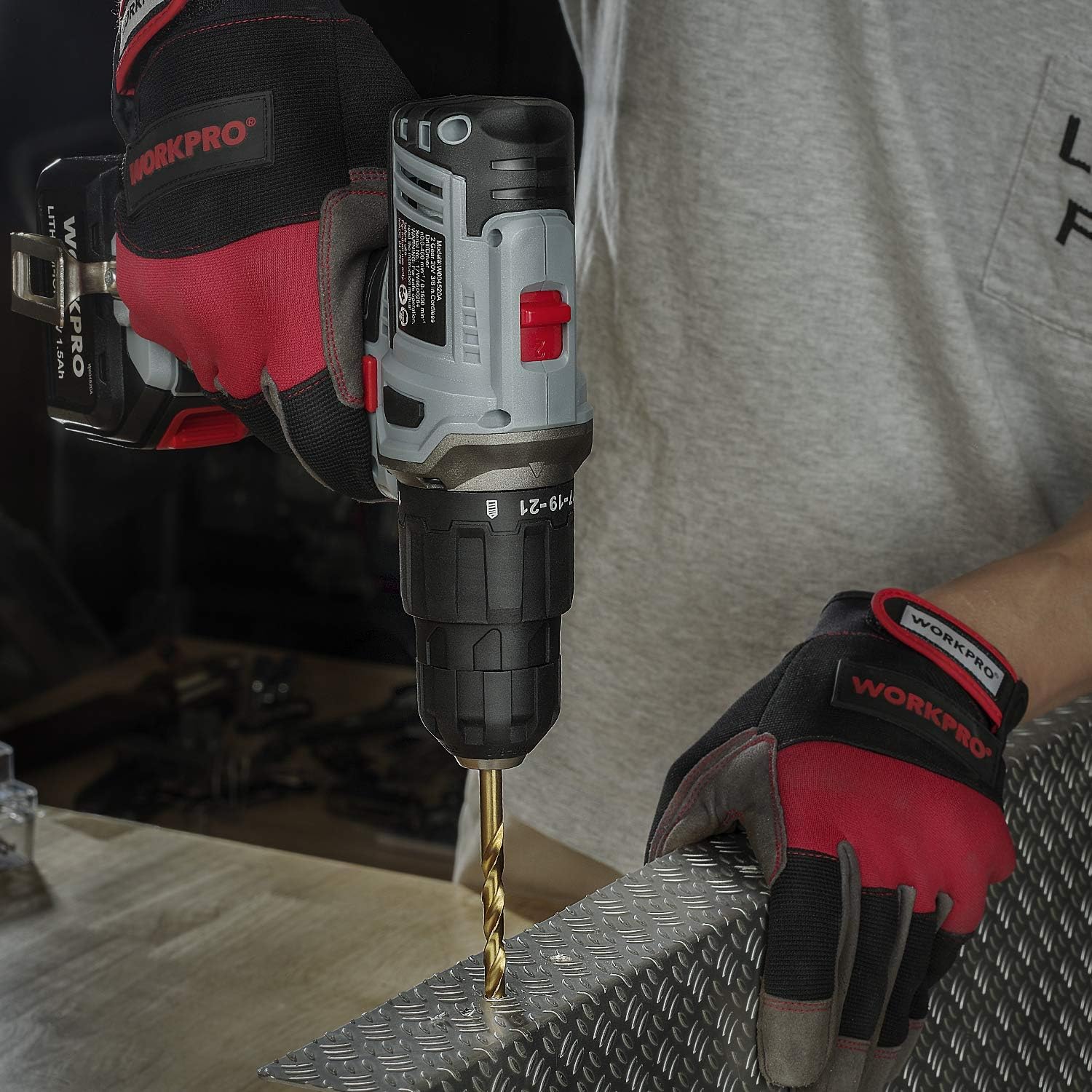Gloved hands using a WORKPRO cordless drill with drill bit on a metal surface