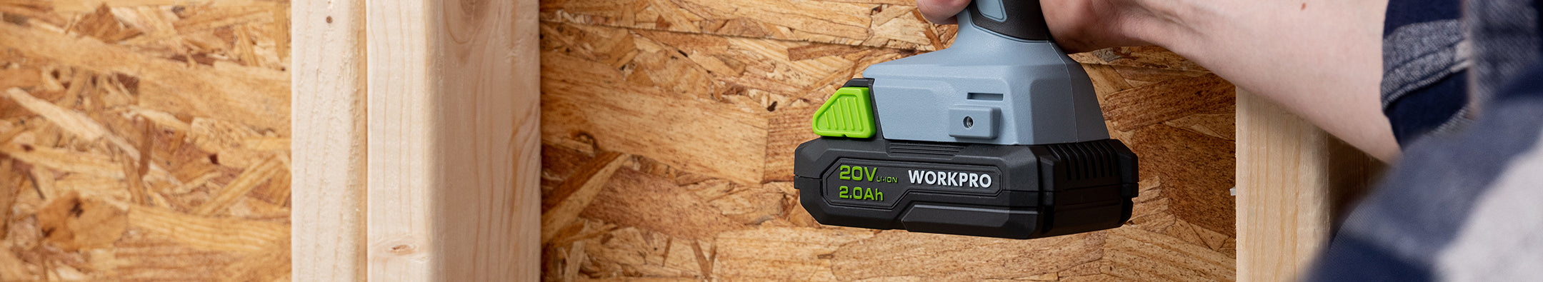 Hand holding a gray and black power tool with green accents, resting against wooden boards in a workshop