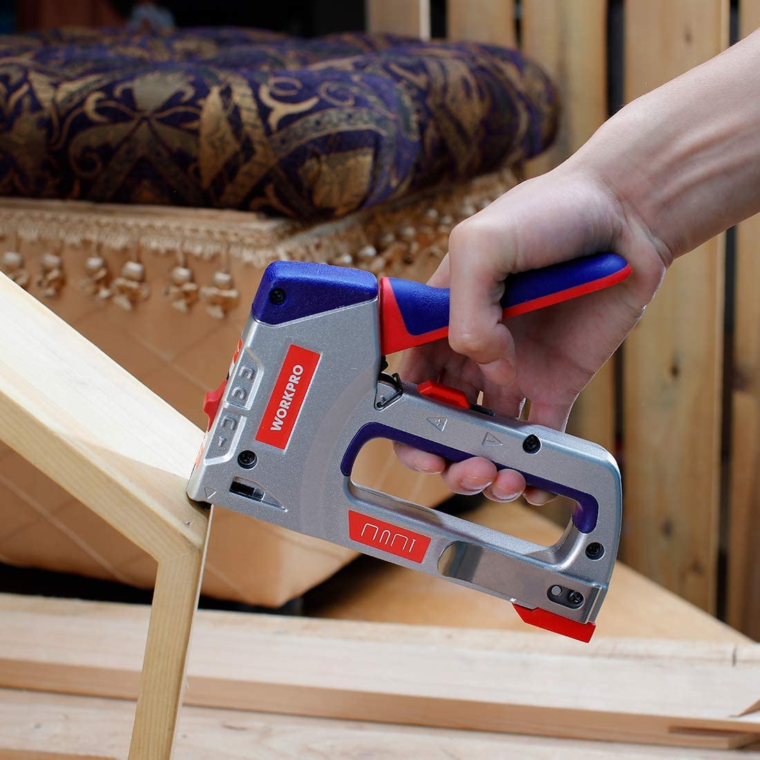 Manual staple gun with blue/red handle firing brads into a wooden frame