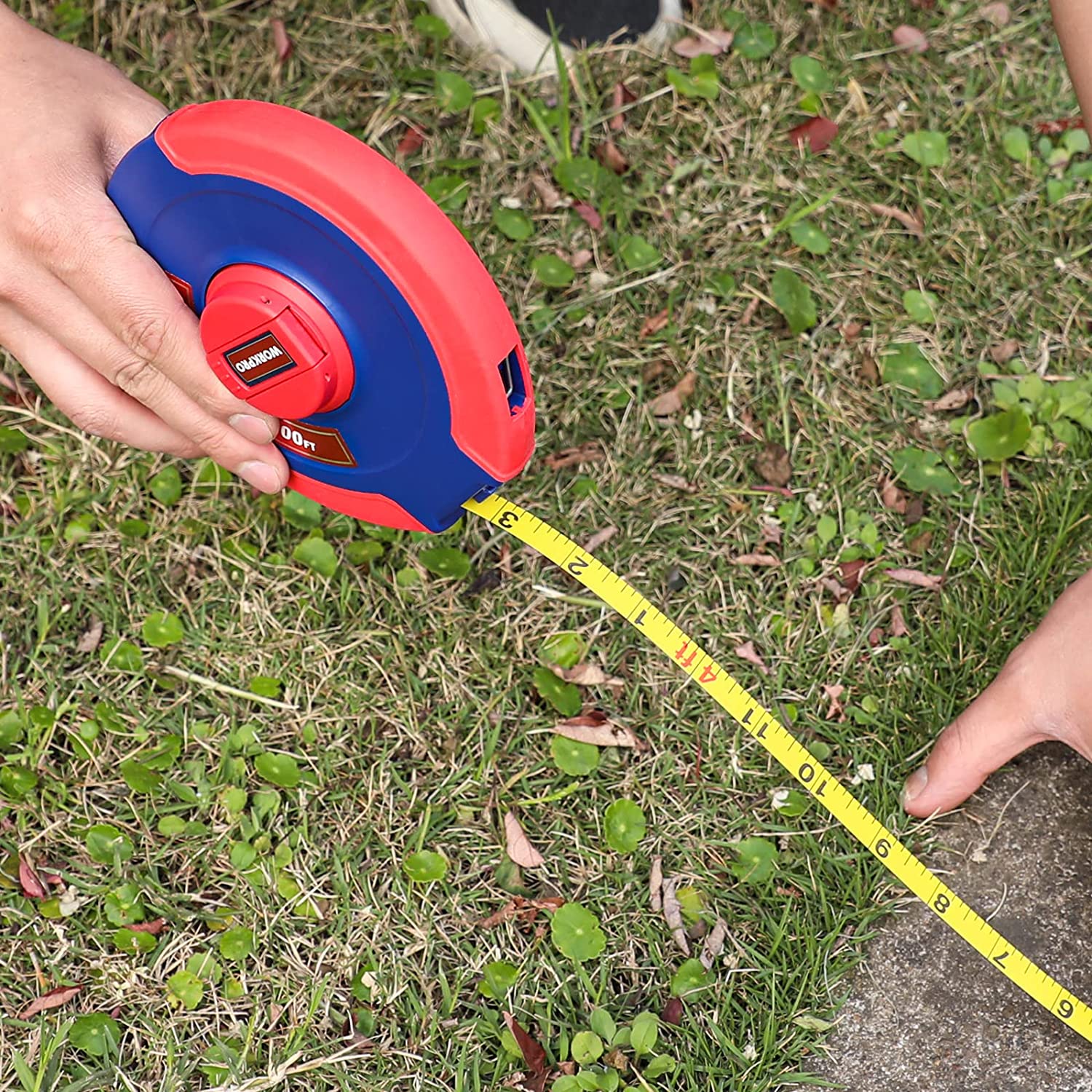 Red and blue tape measure reel laying yellow tape across grass for measuring