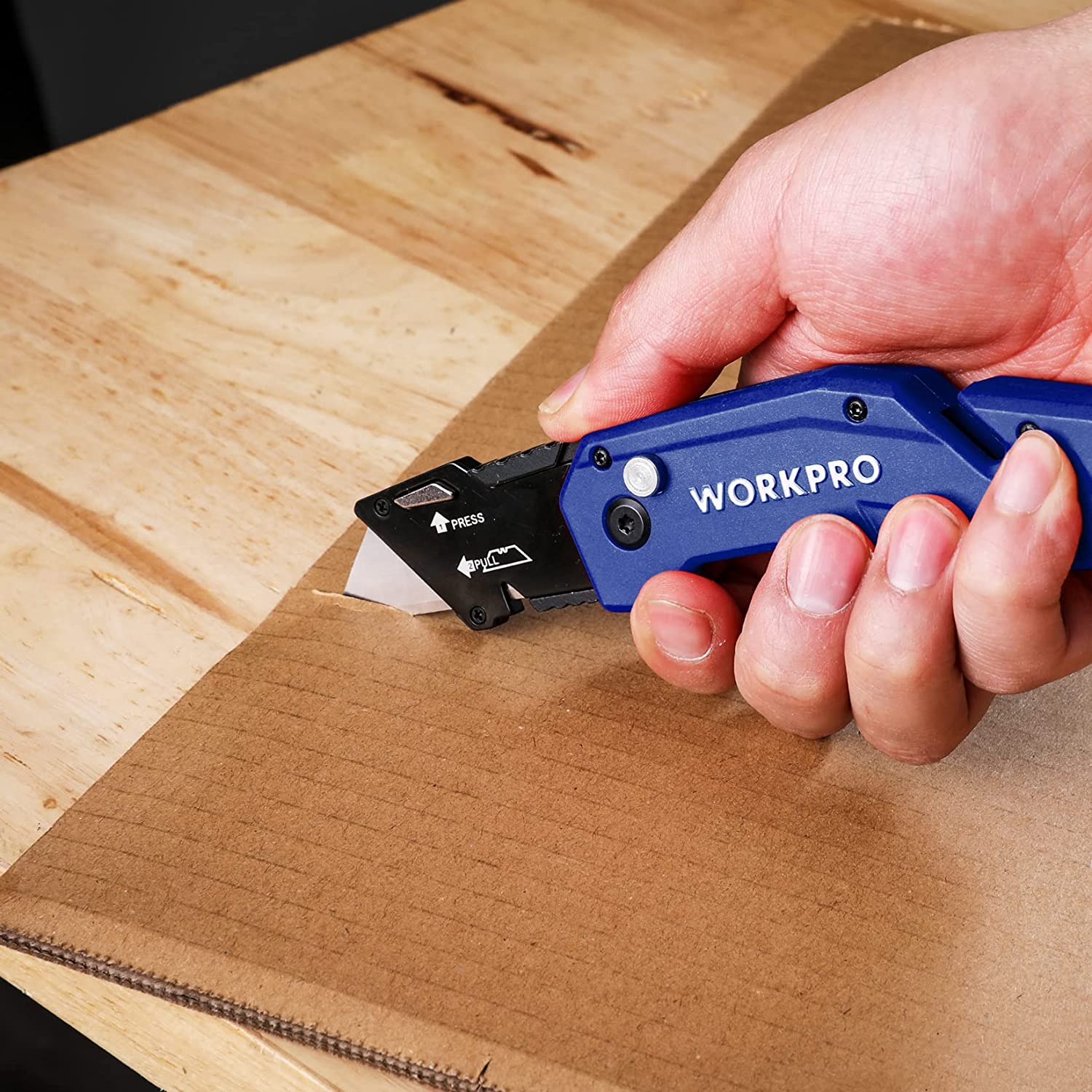 Blue folding utility knife with black blade cutting cardboard on wood board.